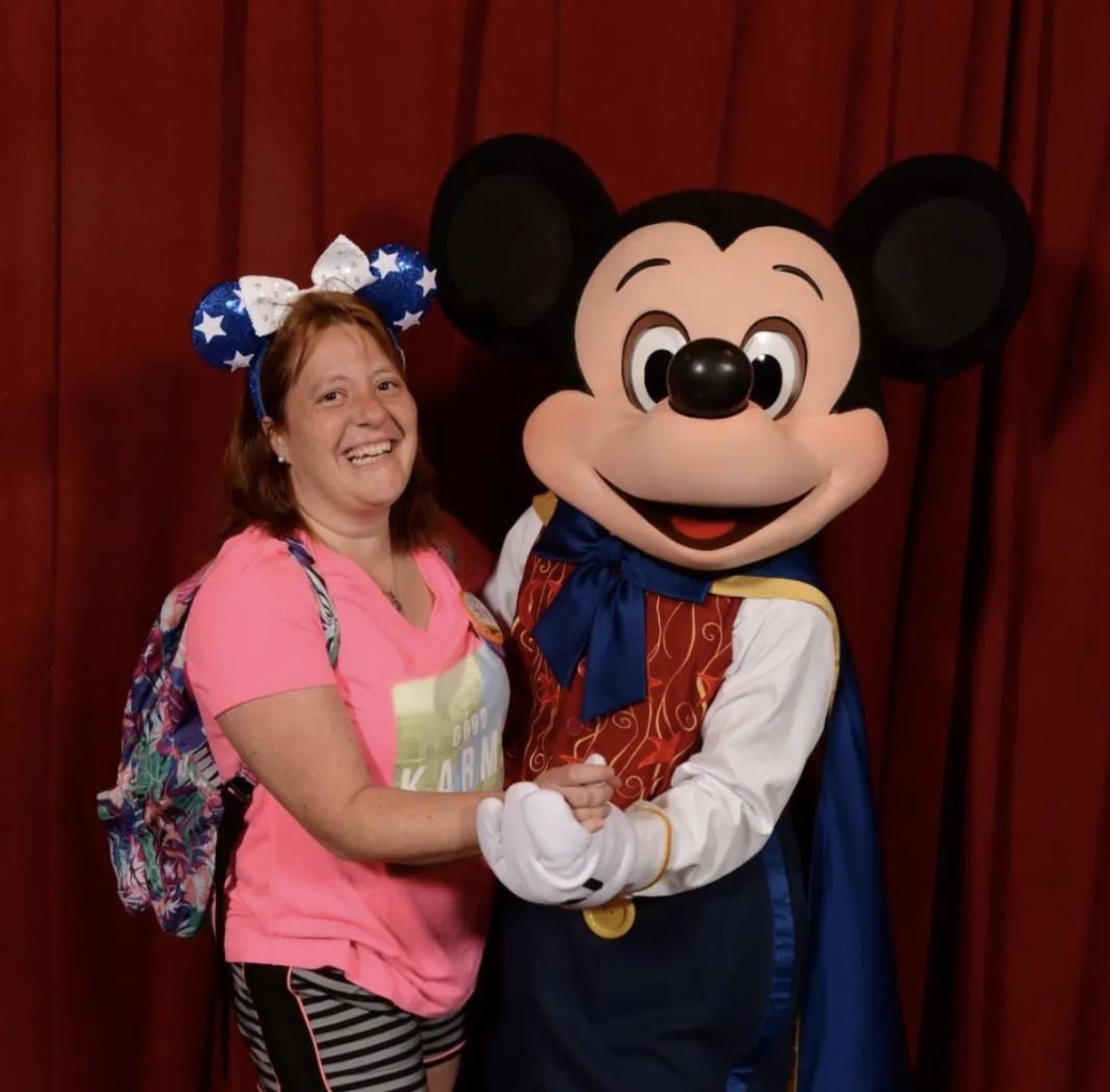 Woman smiling with Mickey Mouse at Disney park meet-and-greet in front of red curtain