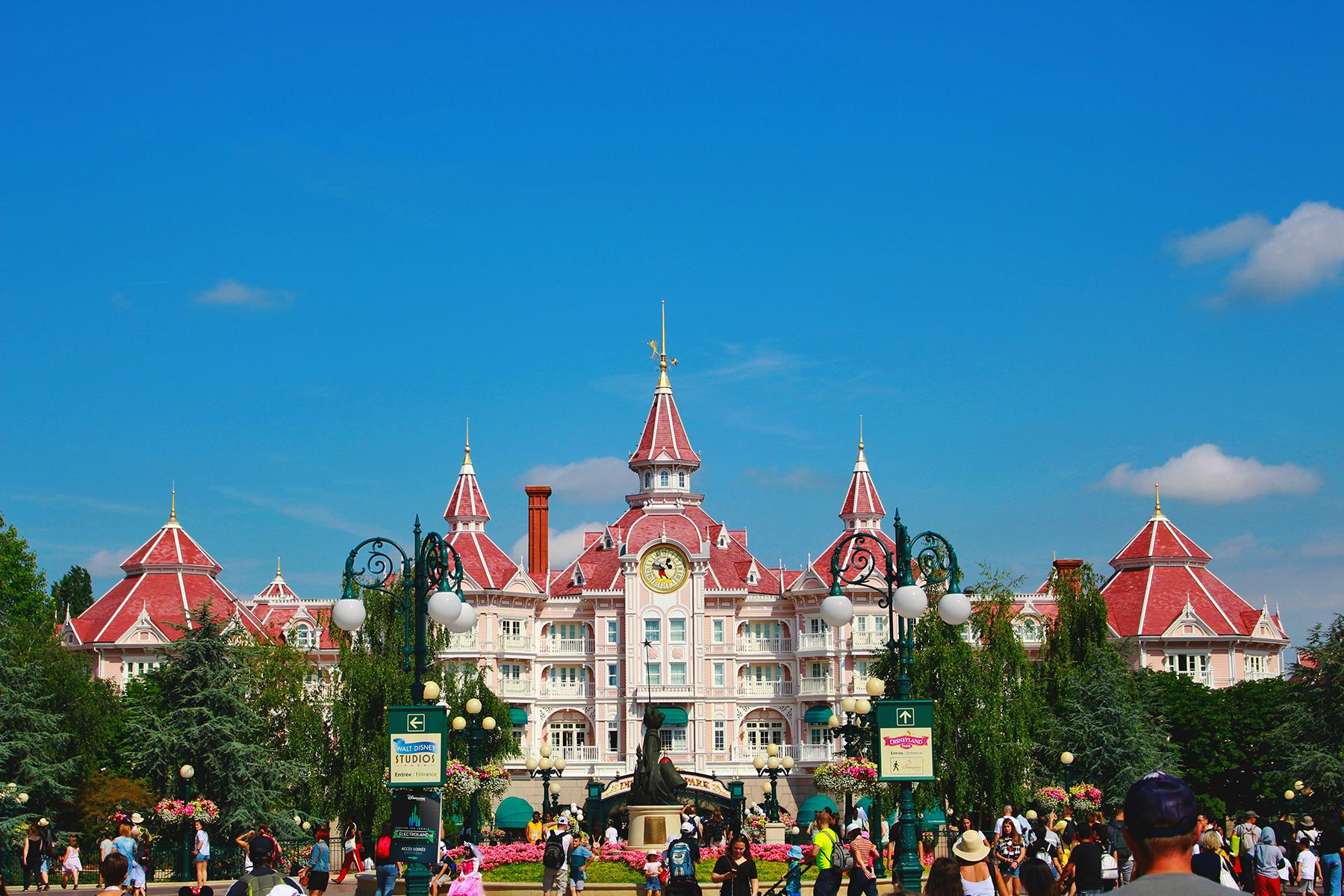 Disneyland Paris castle entrance with red rooftops, clock tower, gardens, and excited visitors.