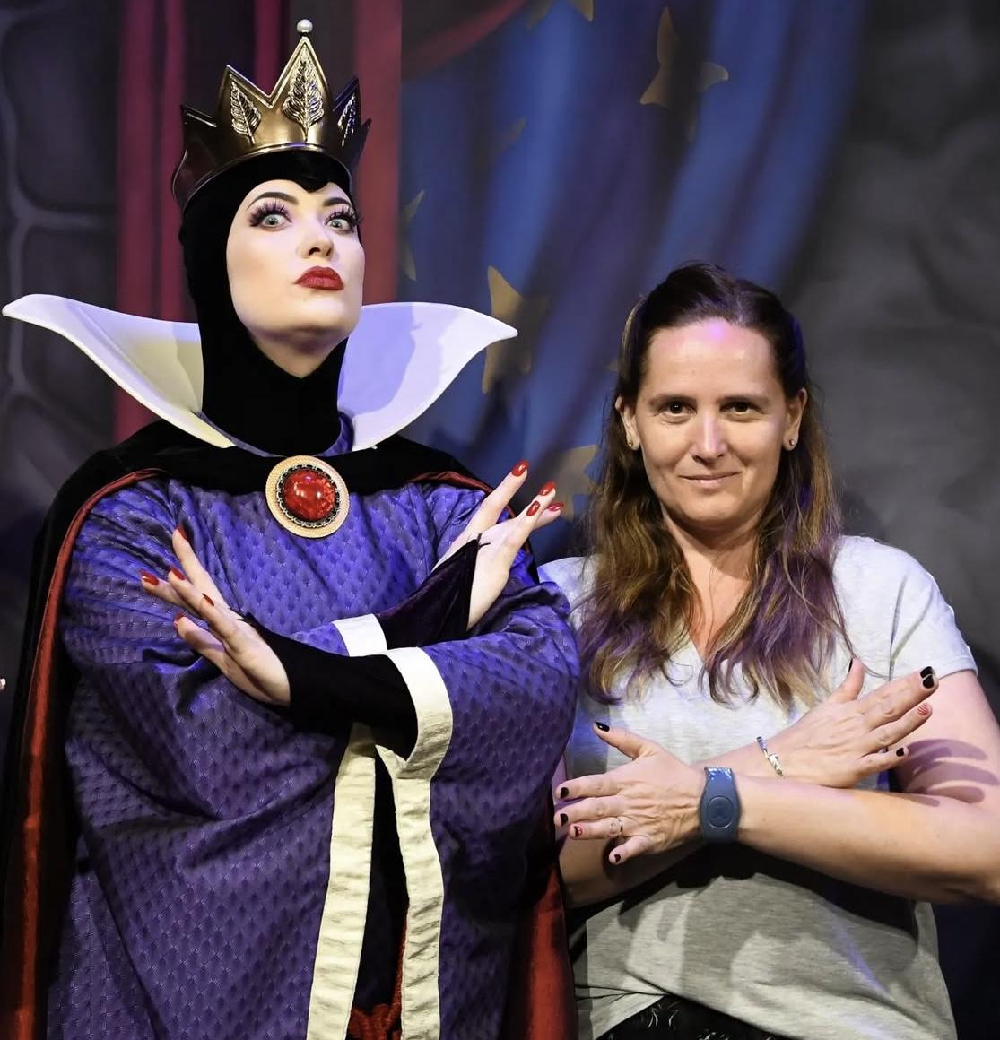 Evil Queen cosplay posing with smiling friend in fairytale-themed photo backdrop.