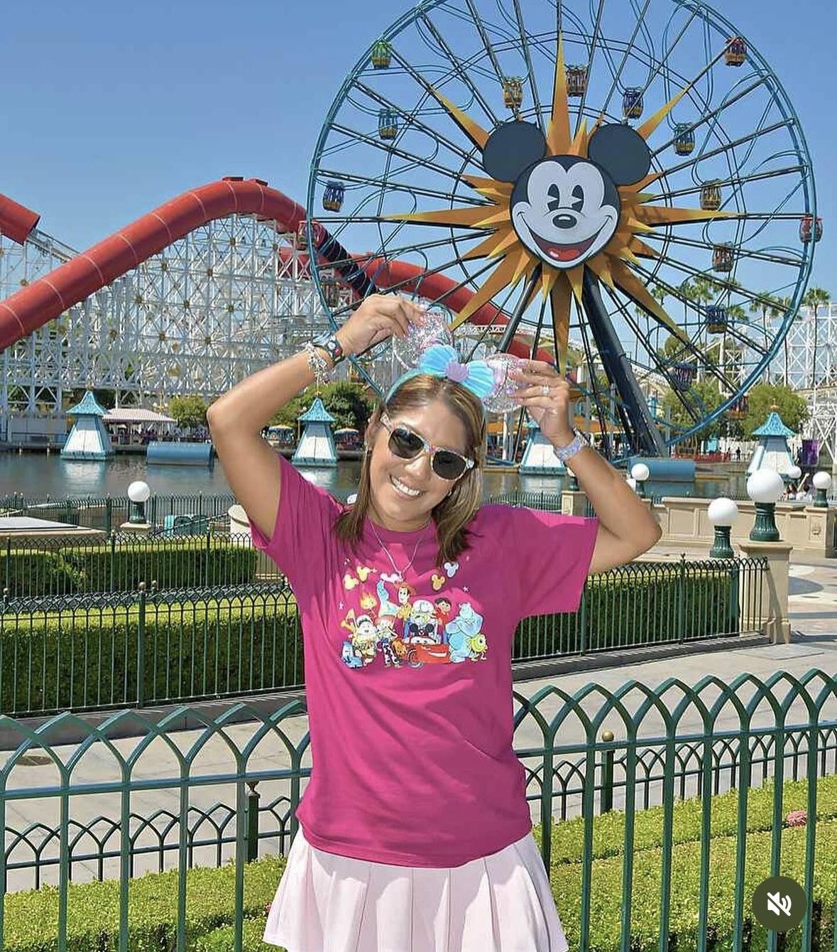 Smiling woman with Mickey ears posing by Ferris wheel at Disney theme park.