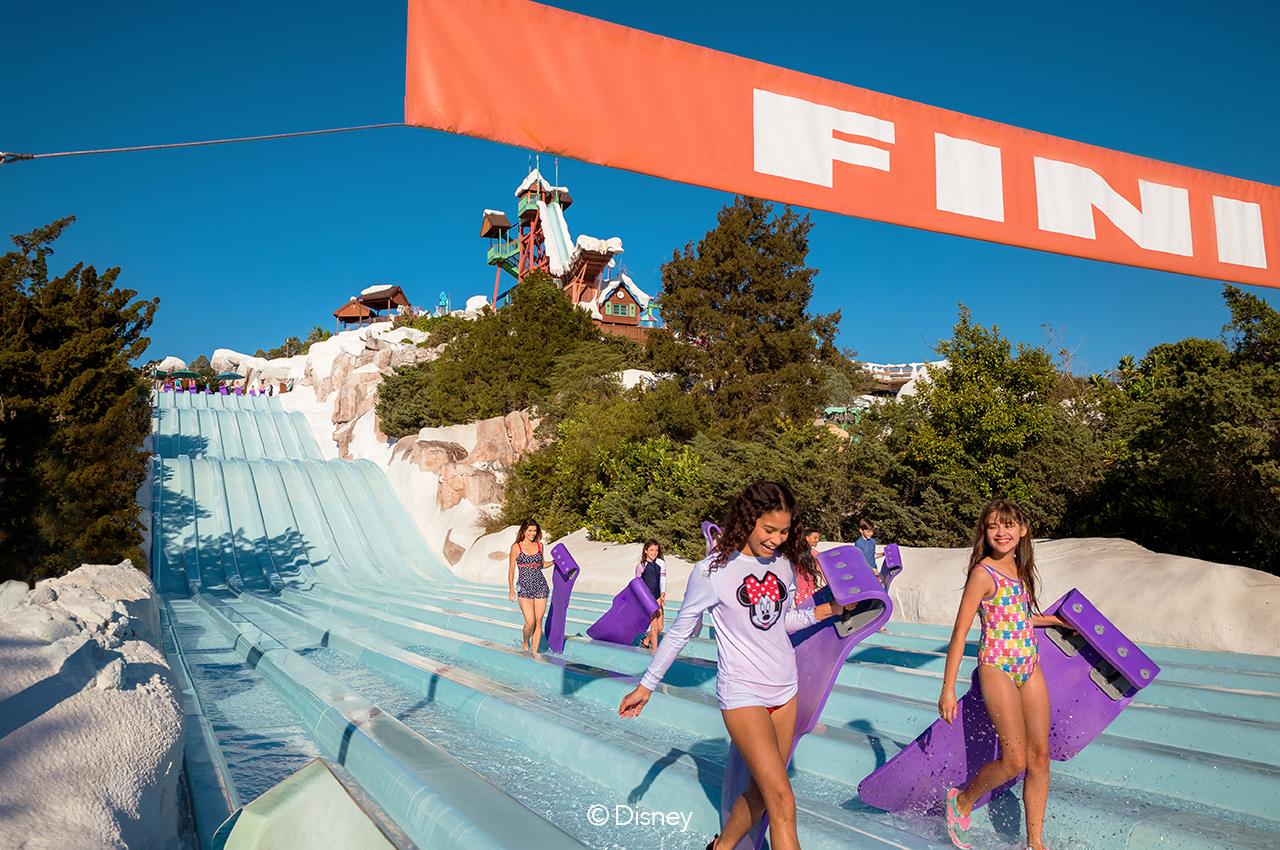 Kids sliding on bright blue water slide with body boards at summer water park.