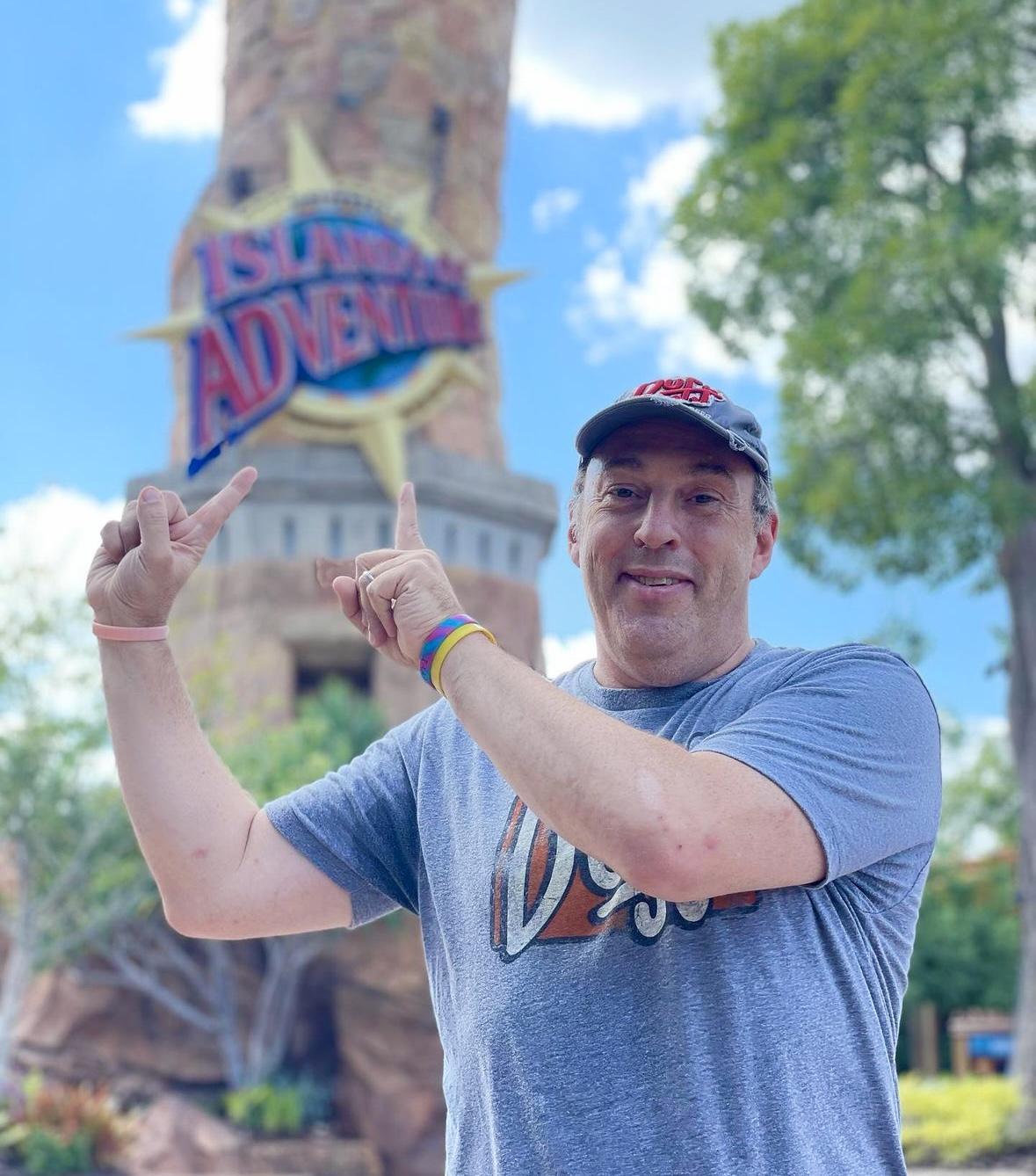 Man pointing happily at Island Adventure theme park sign on a sunny day.