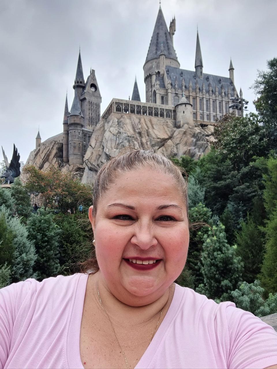 Woman smiling selfie with fantasy castle backdrop and lush greenery in enchanting travel scene.