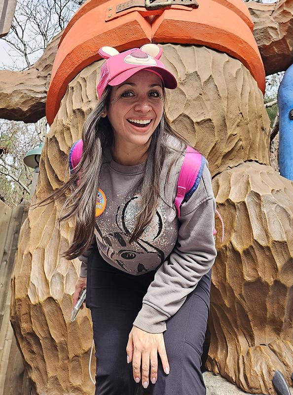 Smiling woman in pink cap poses by whimsical wooden statue outdoors.