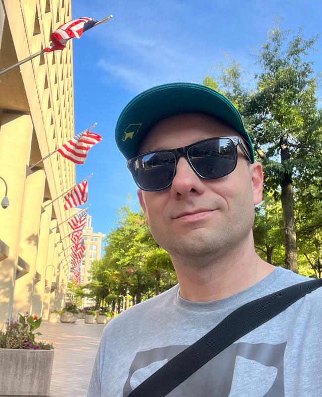 Man in sunglasses and green cap takes selfie with flags, trees, and modern architecture.