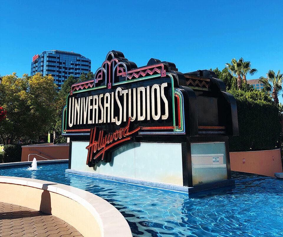Universal Studios Hollywood entrance sign with fountain, colorful art deco design, and clear blue sky.