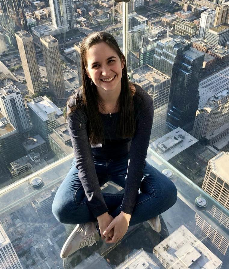 Woman sitting on glass platform above city skyline with fearless smile and urban view.