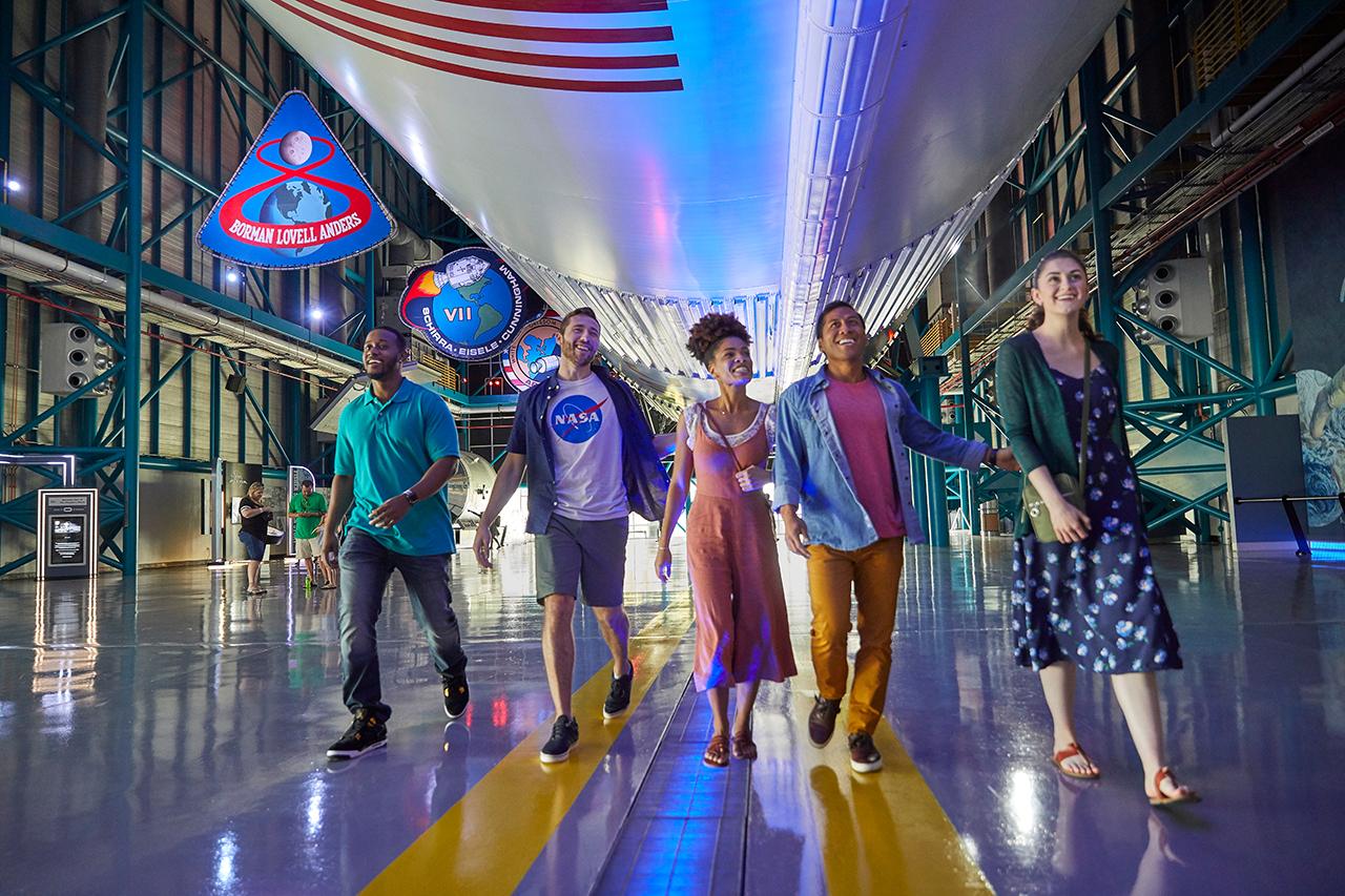 Diverse young visitors exploring NASA Kennedy Space Center with spacecraft and American flag backdrop