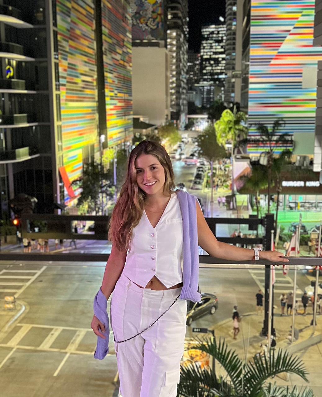 Stylish young woman smiling on city balcony with colorful nightlife skyline.