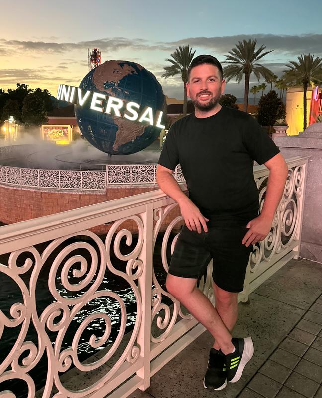 Man posing by Universal Studios globe at sunset with palm trees and colorful skies.
