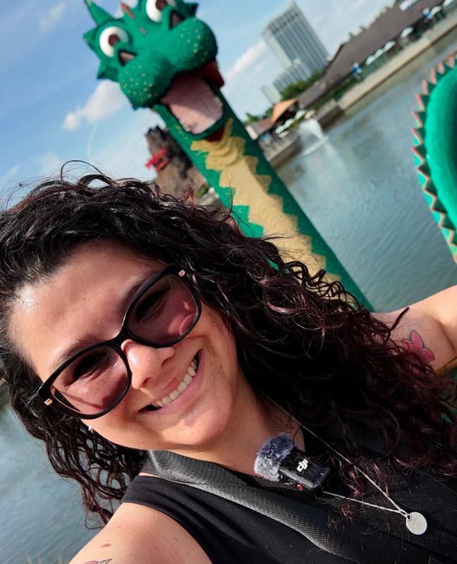 Smiling woman selfie with green dragon statue by the water on a sunny day.