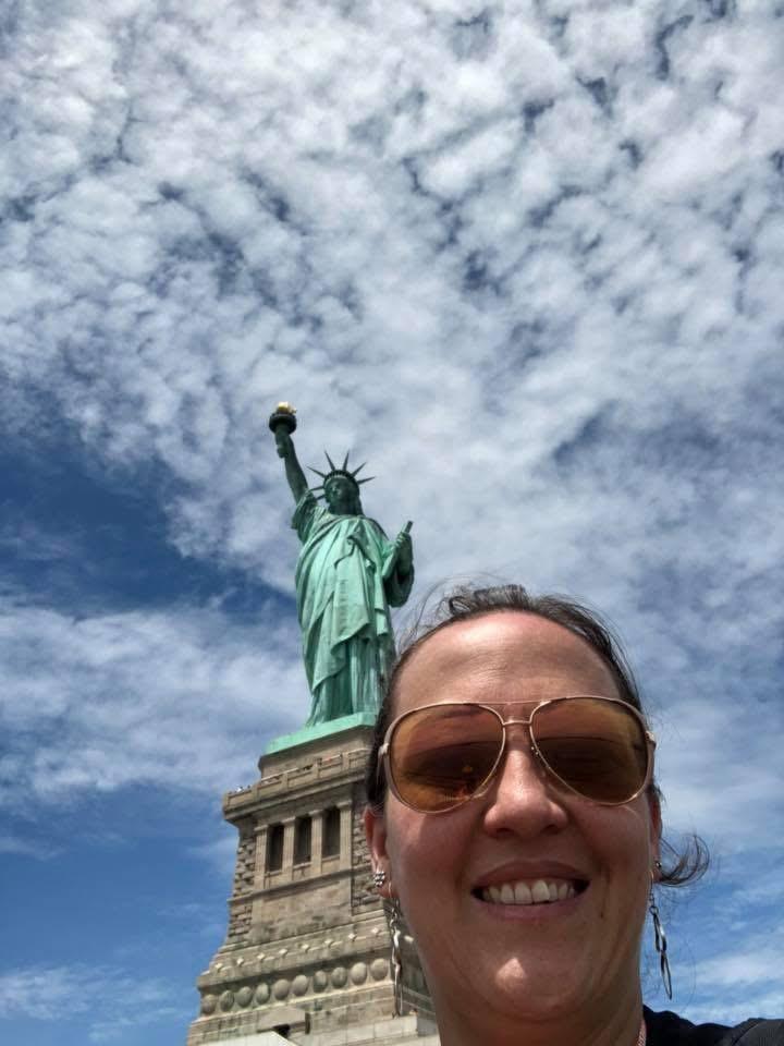 Woman smiling selfie with Statue of Liberty under bright blue sky in New York.