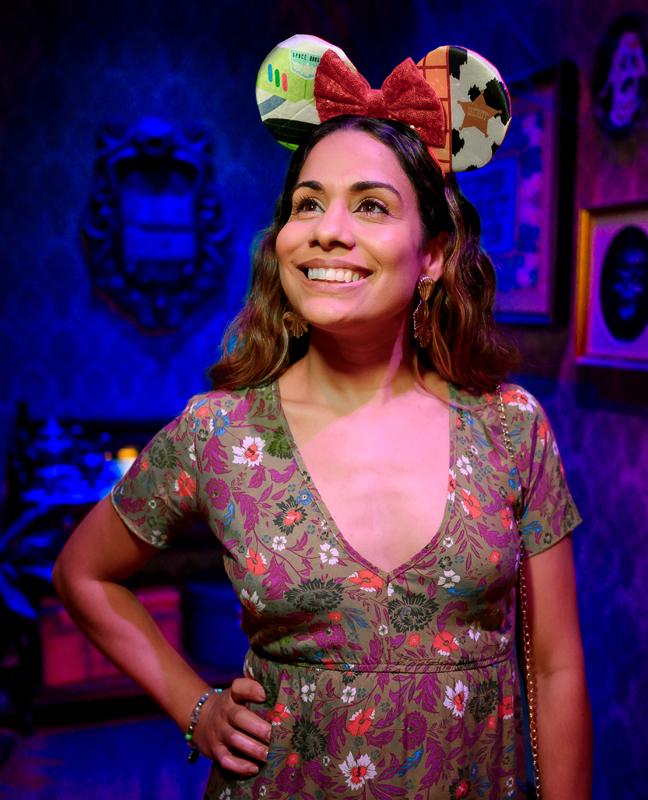 Smiling woman in floral dress with colorful mouse ears at vibrant theme park.